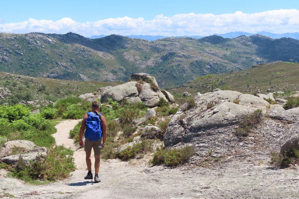 Man in shorts with blue backpack walking down rocky road with green mountains in background under blue sky.