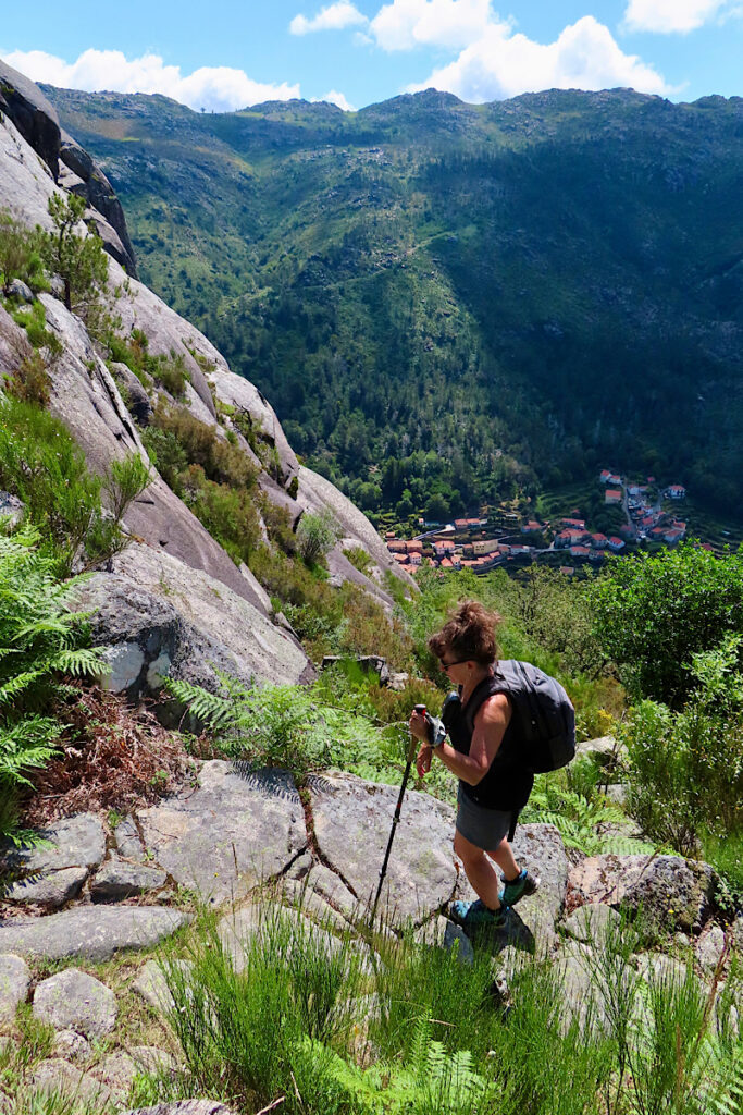 Woman in short sleeve shirt and shorts wearing a backpack climbing rocky trail using hiking poles in mountains terrain.