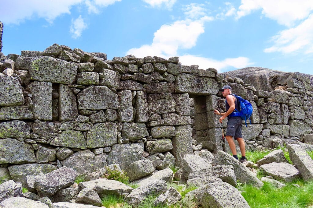 Man in shorts with blue backpack entering doorway of ruined stone building.