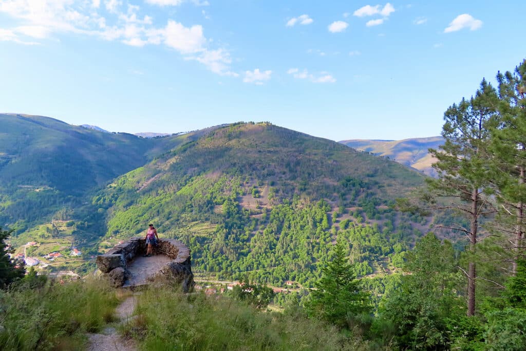 Woman in pink shirt and blue shorts standing on lookout platform with forested slopes behind her.