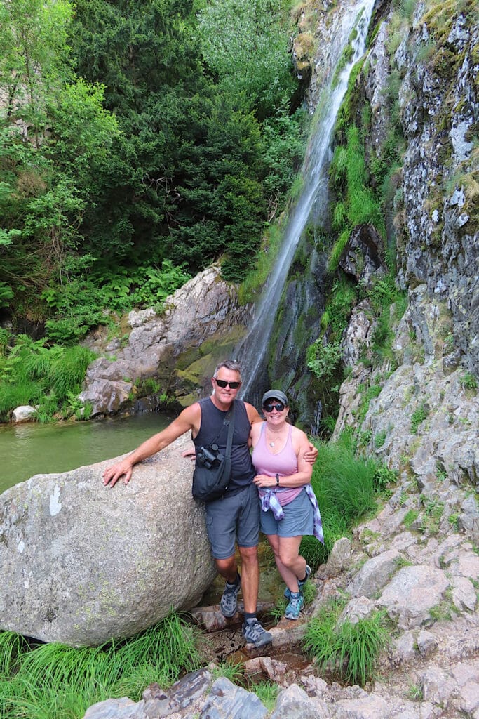 Man in black shirt and shorts standing beside woman in pink shirt and blue shorts with waterfall in background.