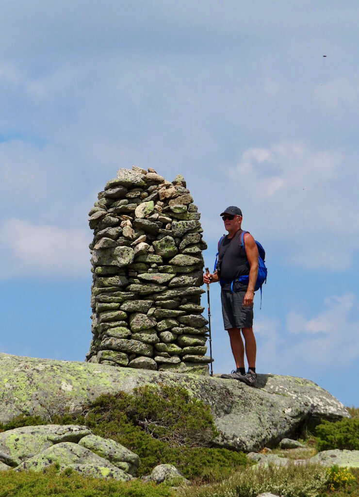 Man in short wearing backpack standing near to tall rock cairn.