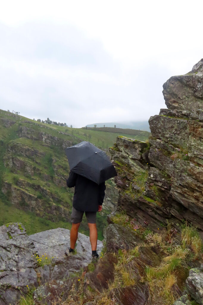 Man in black raincoat and shorts standing under umbrella looking out over a rugged valley.