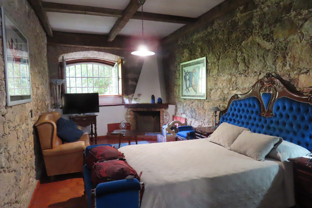 Rustic stone-walled room and bed with blue velvet headboard and footstool.