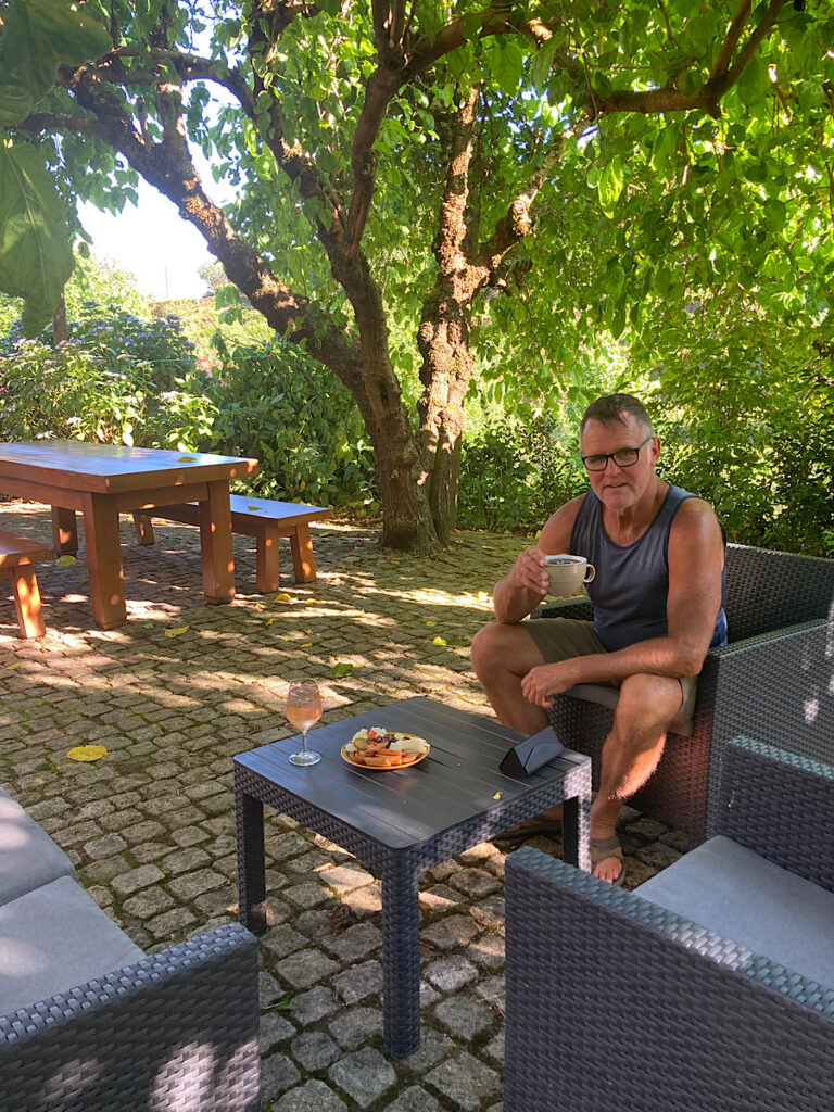 Man holding cup with cold drink while sitting on black patio furniture with green trees in background.