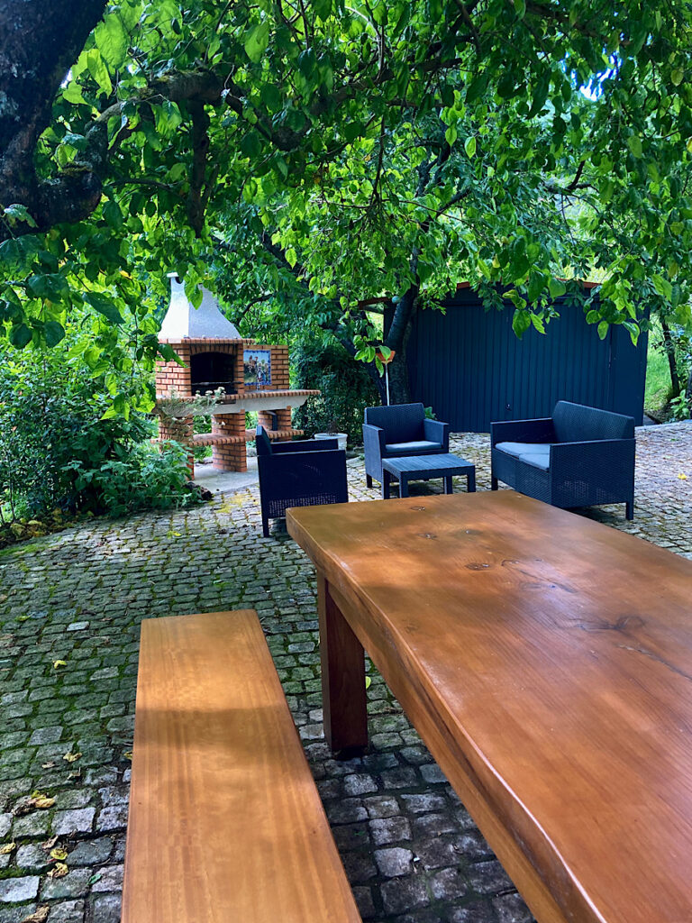 Large wooden table and bench with black conversation table and chair set and barbecue in background.