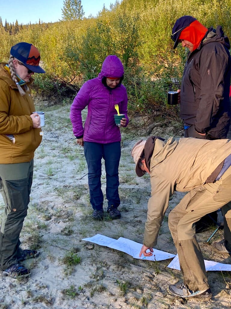 Two woman and one man holding coffee cups looking at a second man bent over a series of paper map pages laid out on sand.