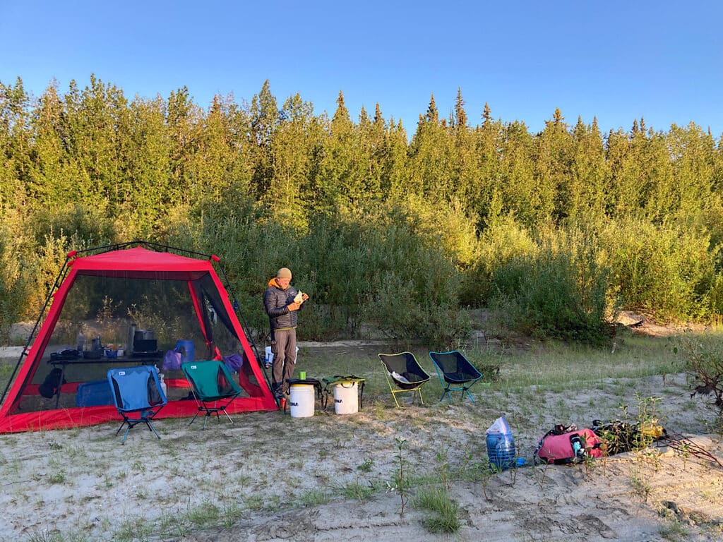 Man in coat and long pants standing beside red bug tent with folding chairs and dry bags around edge.