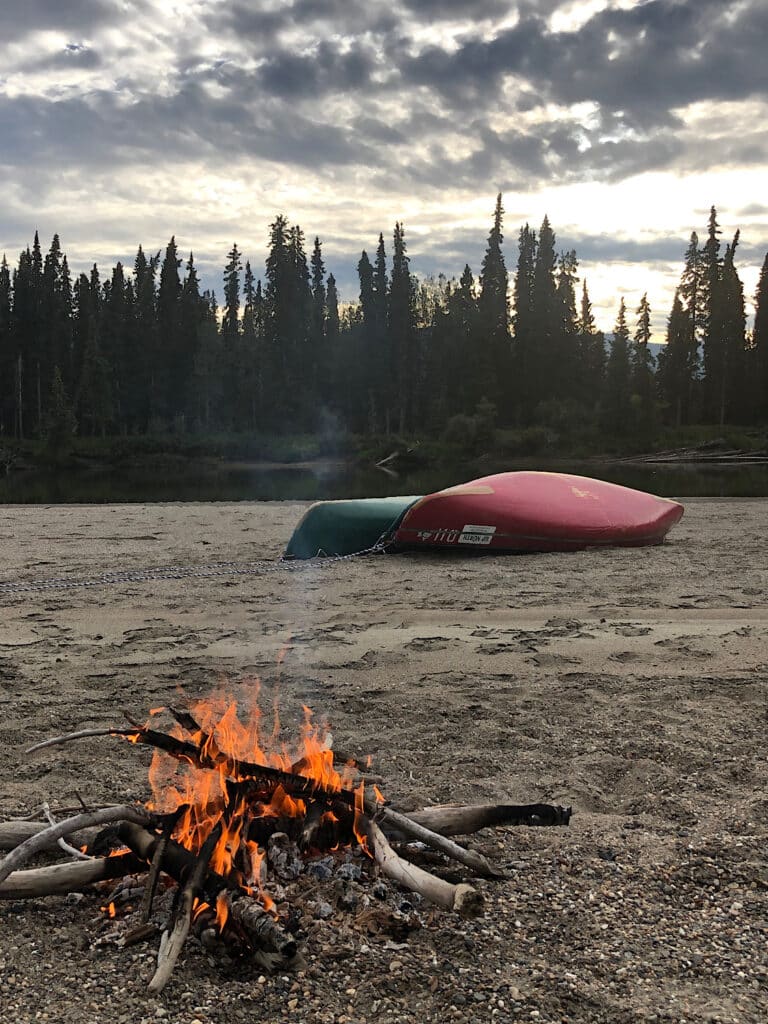 Green and red canoes stacked upside down behind small campfire on sand.