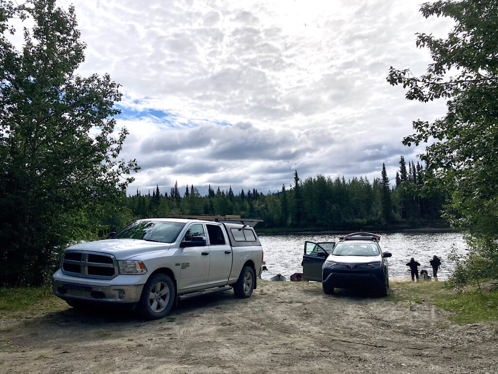 White pickup and dark grey car parked by opening in trees along a river.