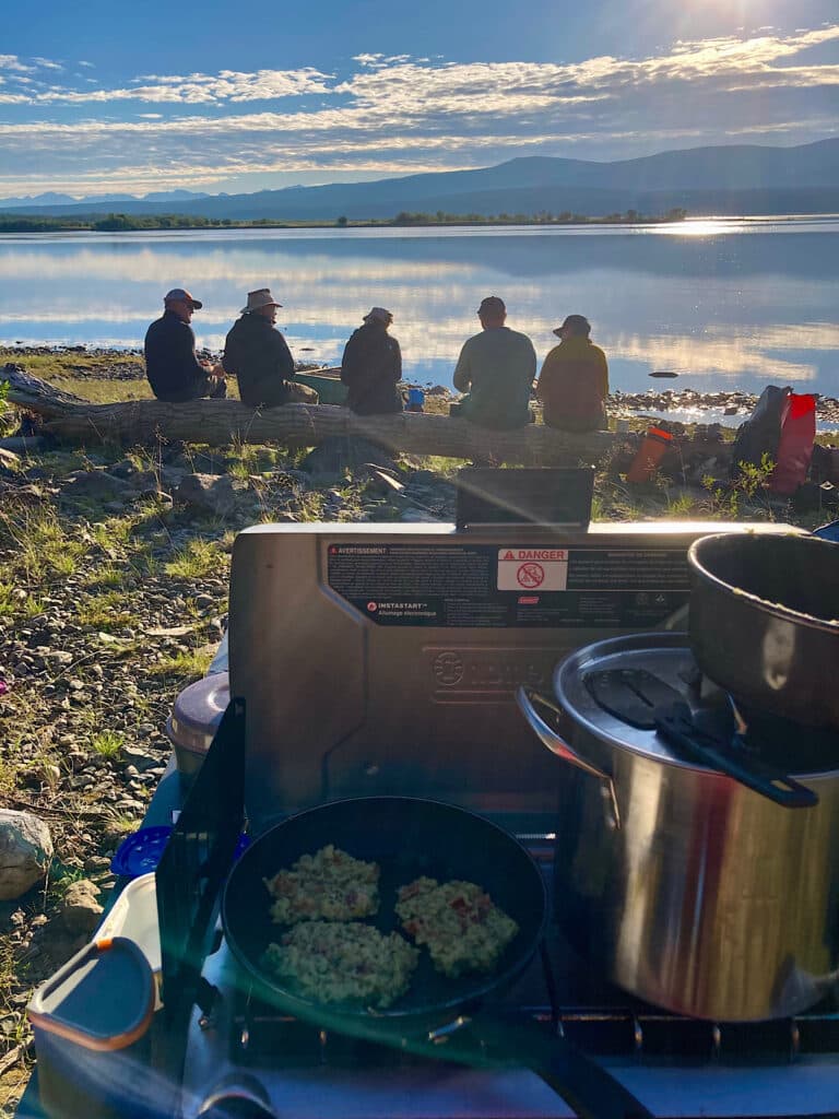 Five people sitting on log in background facing a lake with camp stove set up in foreground.