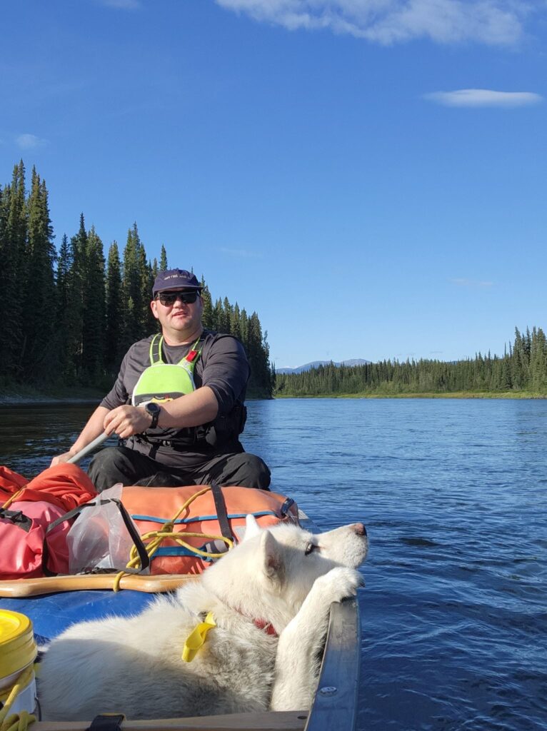 Man paddling full canoe on river with white dog resting chin and paw on gunnel.