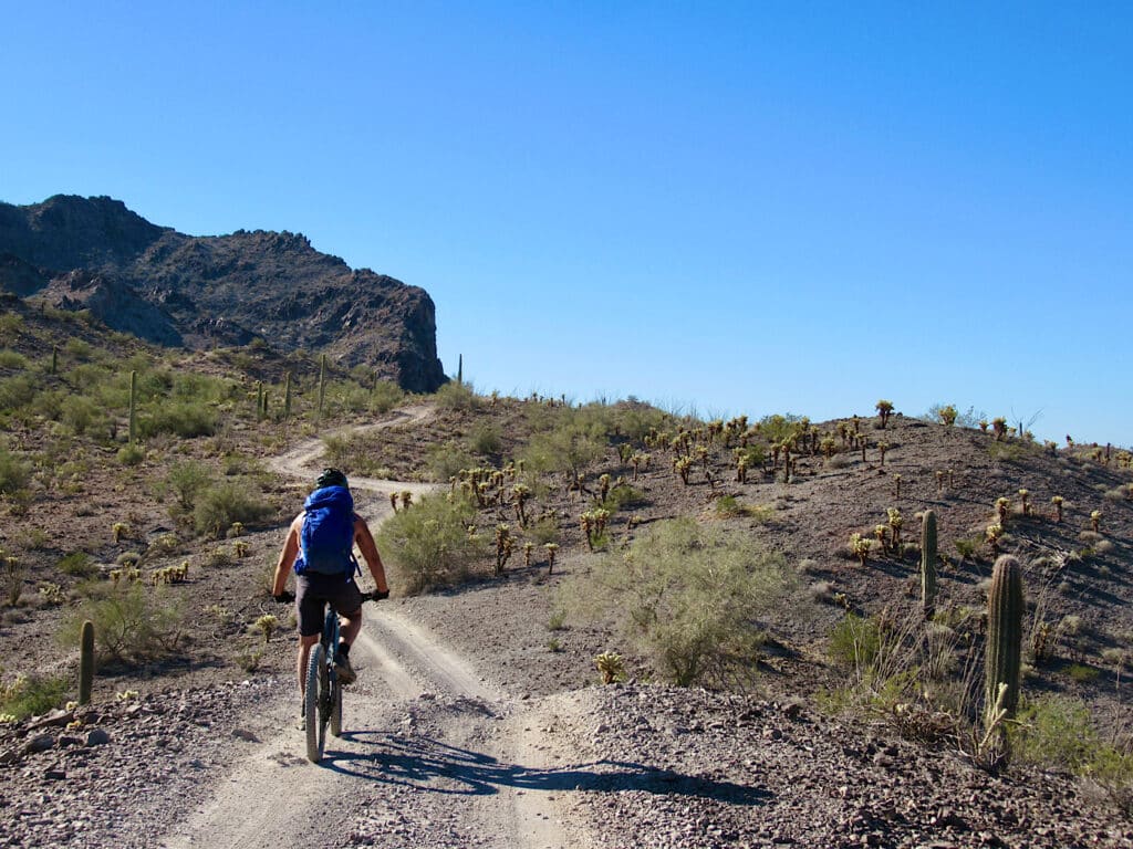 Man riding mountain bike on narrow dirt road with dark mountain in background under blue sky.