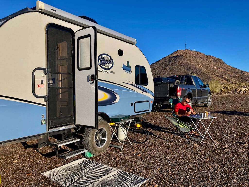 Man in red shirt and shorts sitting in camp chair outside of a blue and white trailer.