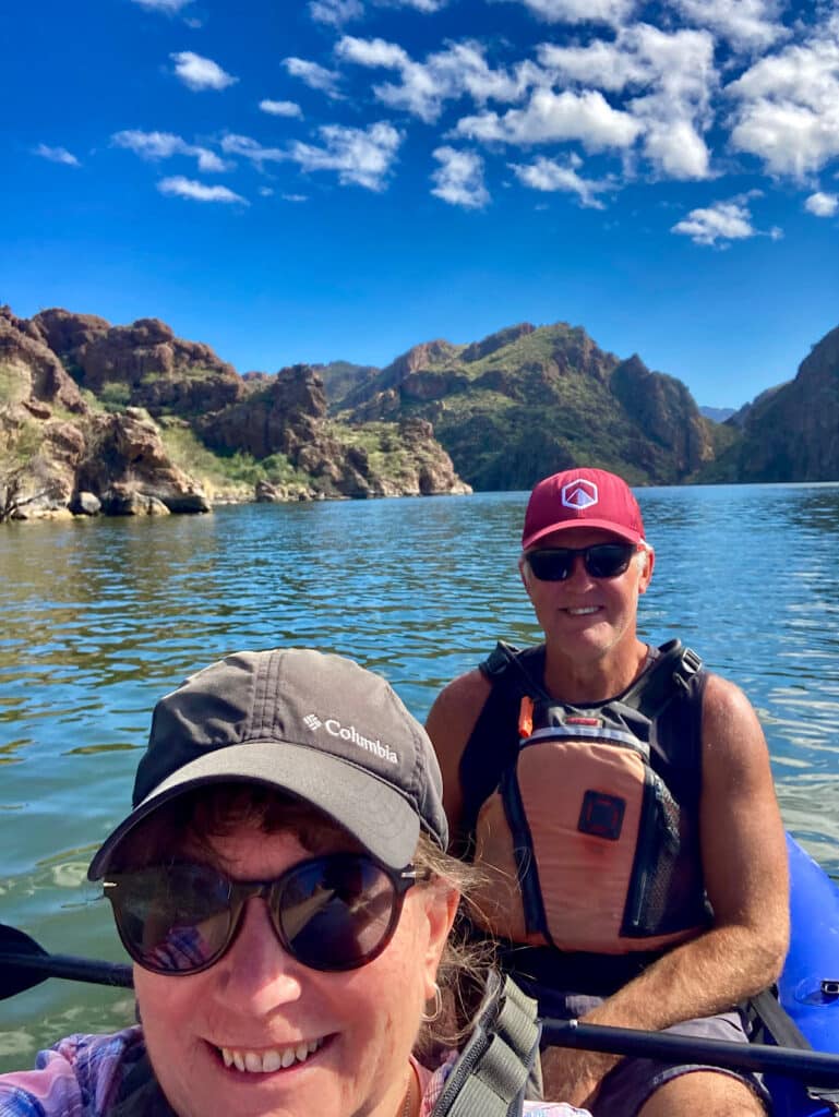 Man and woman smiling at camera while seated in an inflatable kayak on a lake with rugged mountains in background.