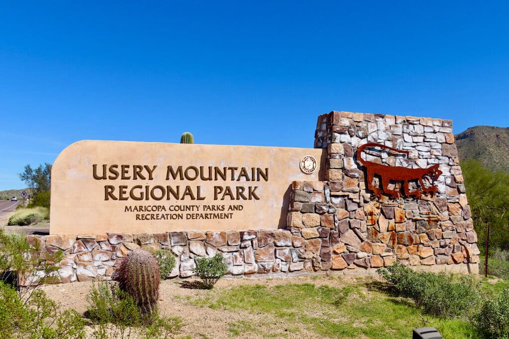 Entrance sign made of rock and pinkish stucco with words "Usery Mountain Regional Park" and metal cutout of a rock art image of a mountain lion.