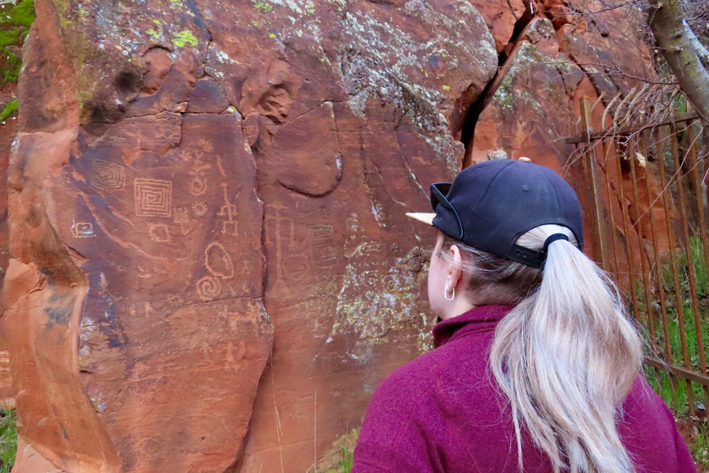 Woman with blonde hair under a black ball cap and wearing a purple jacket looking at a red wall of rock art.