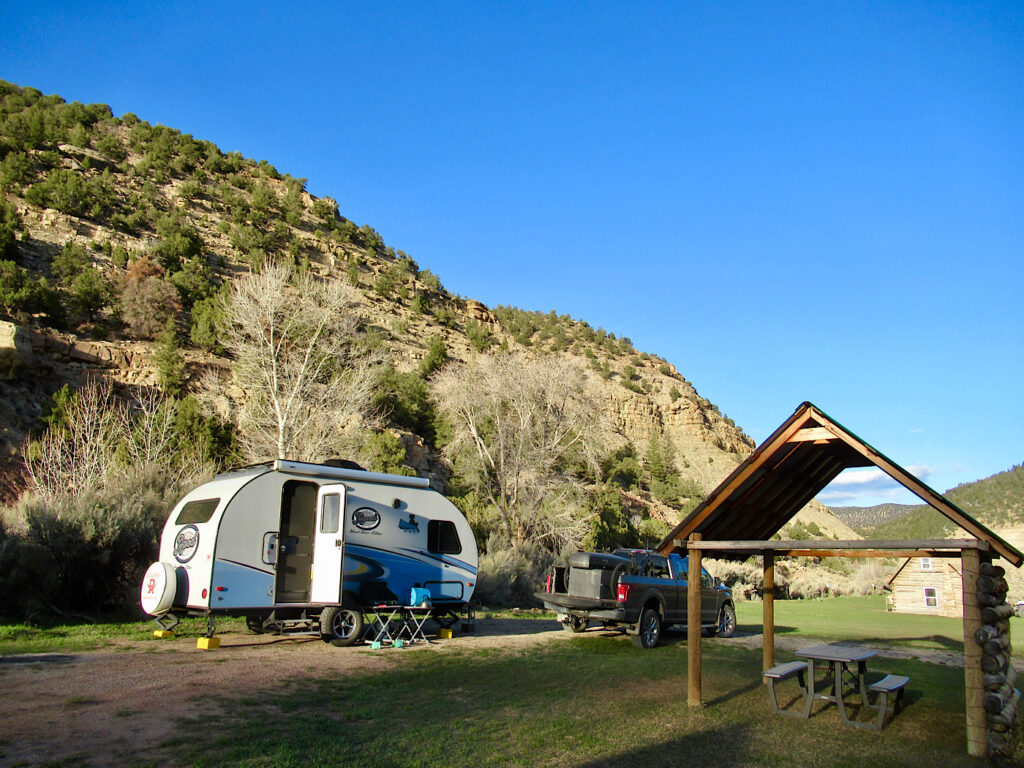 Blue and white trailer set up in campground surrounded by low cliffs under blue sky.