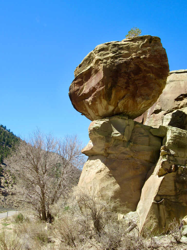 Large boulder balance precariously on smaller column of rock beside road under blue sky. 