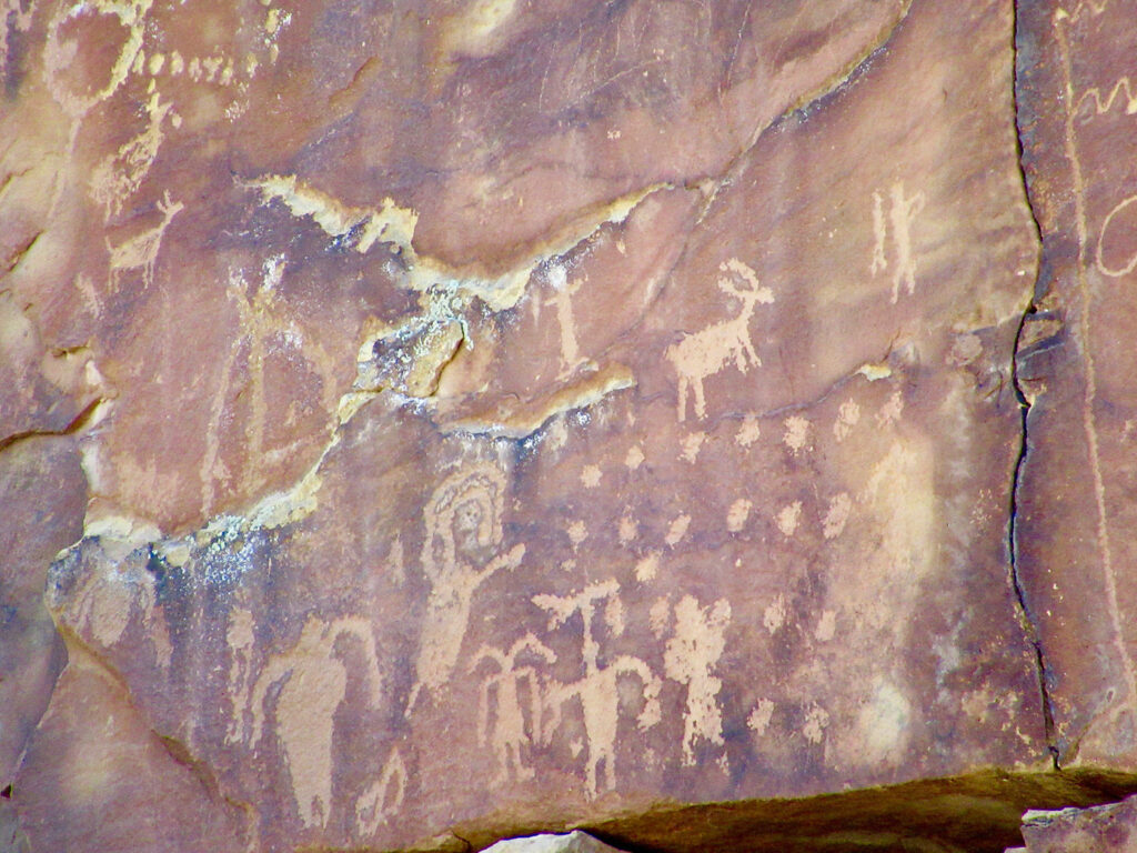 A variety of anthropomorphic figures and a sheep along with rows of dots pecked into red sandstone.