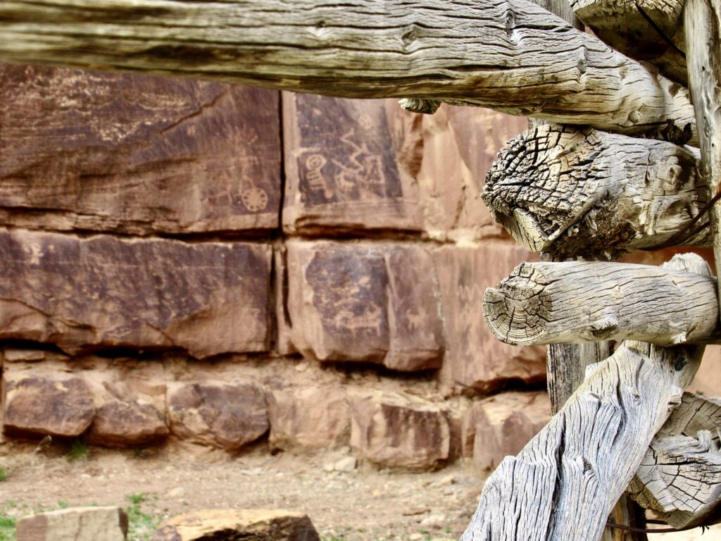 Rock art pecked into red sandstone wall behind weathered grey log fence.