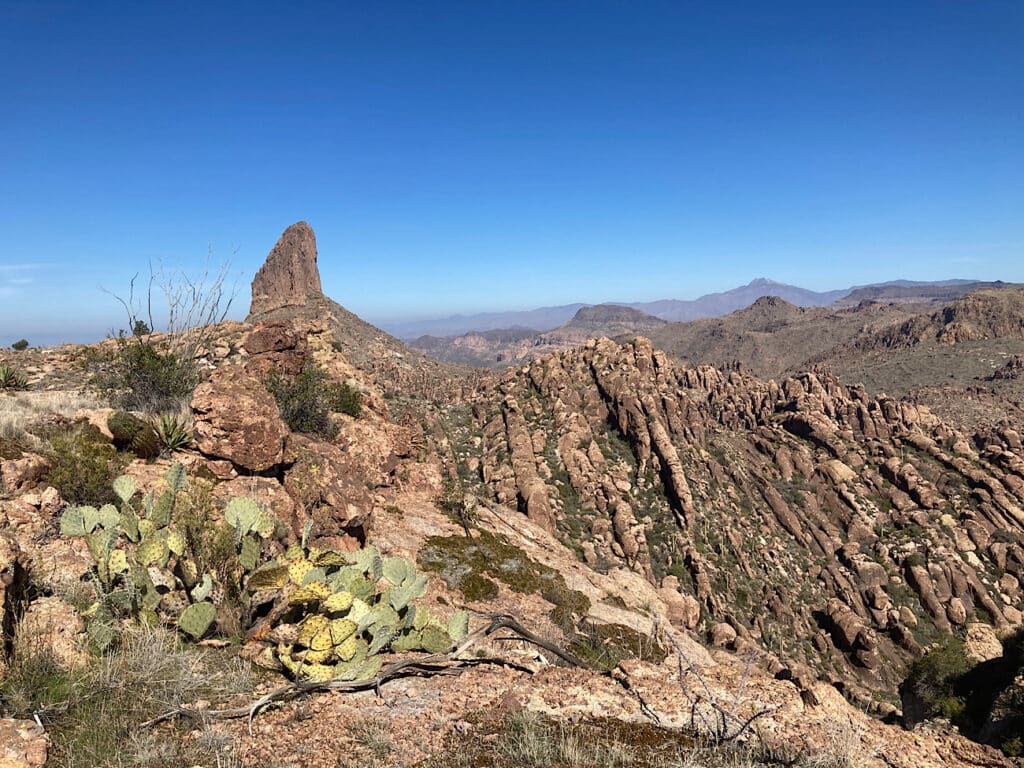 Rugged brown rocky terrain with tall pointy peak in distance under blue sky.
