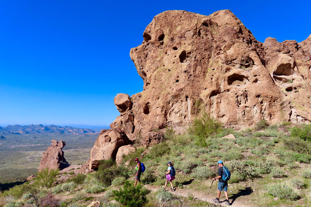 Two men and a women all with backpacks walks trail beside tall, pock-marked red fin of rock.