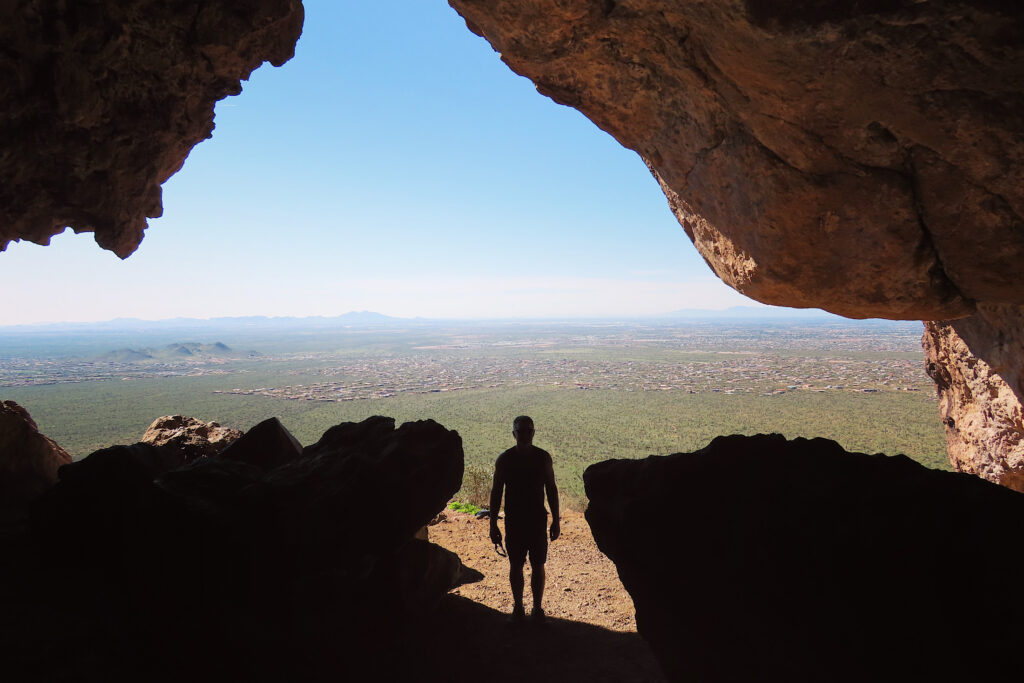 Silouette of man at front of a cave above green valley.