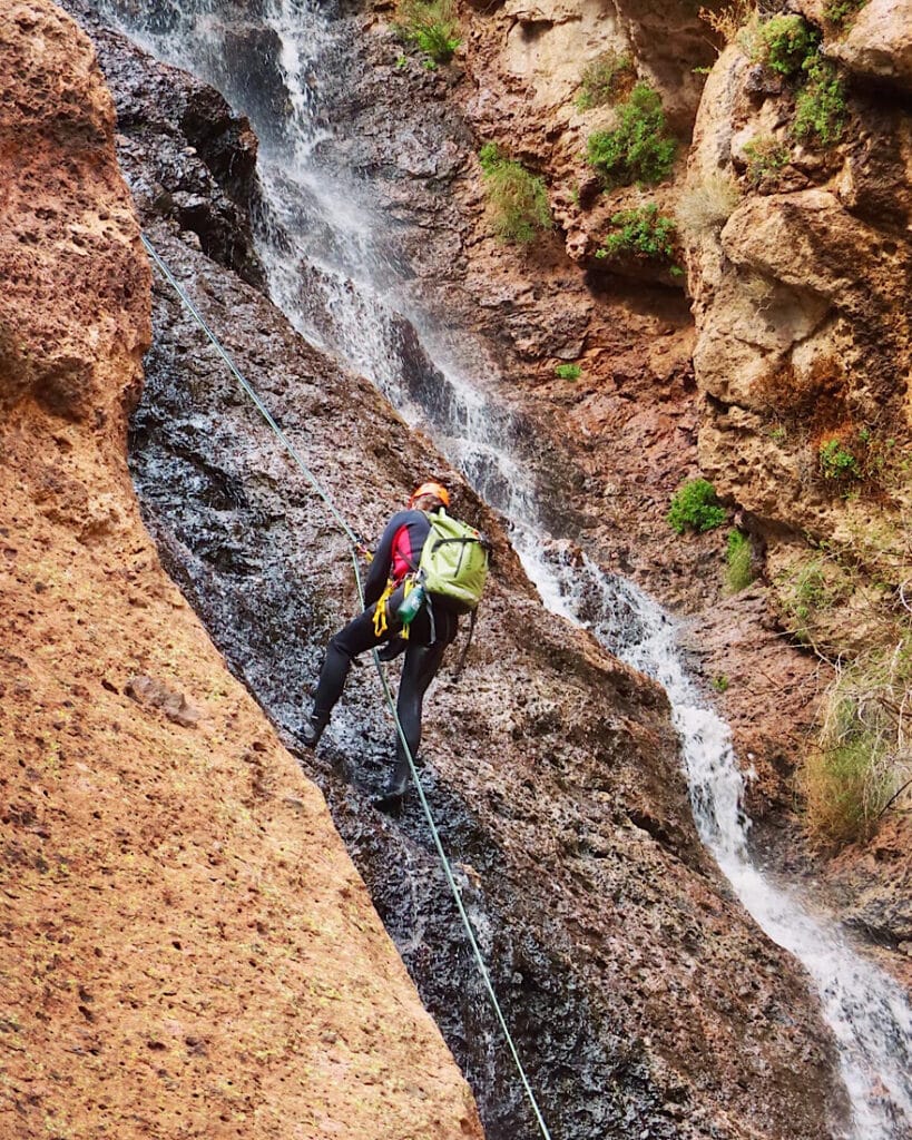 Man in black with bright green backpack on rope in middle of a thin waterfall.