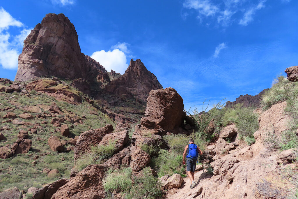 Man in shorts with bright blue backpack walk up rocky trail towards mountain peak in desert environment.