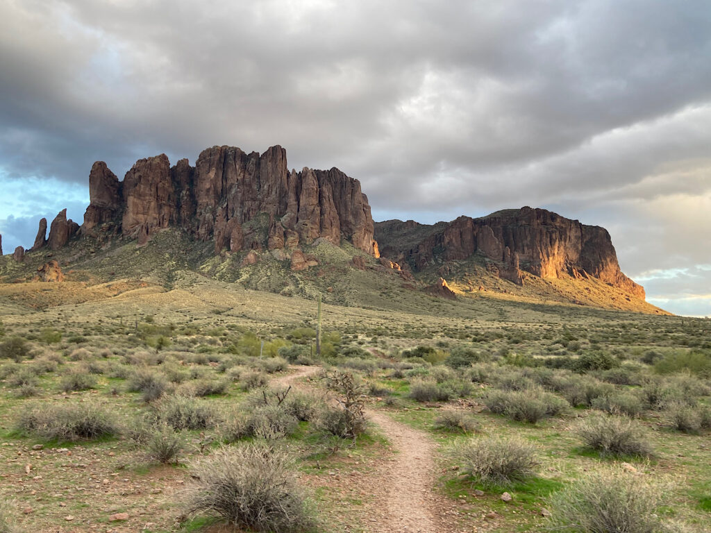 Narrow dirt path in middle of grasses with distant rocky peak under partially cloudy sky.