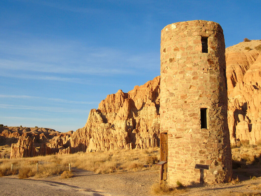 Stone tower with 3 narrow windows in evening glow in front of cliffs under blue sky.