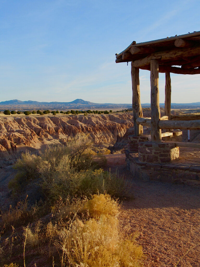 A wood and stone shelter on the edge of a cliff overlooking eroded cliffs under blue sky.