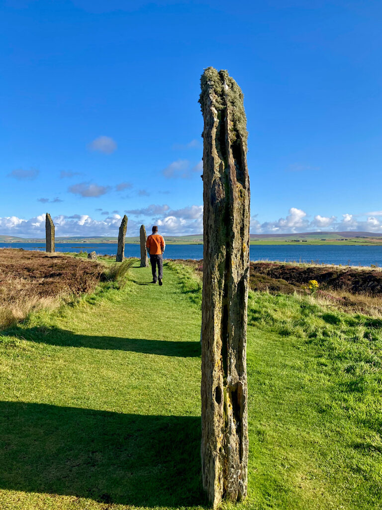 A tall thin slab of rock in the foreground with a man in an orange jacket walking on a grassy burm towards more standing stones next to water under blue sky.