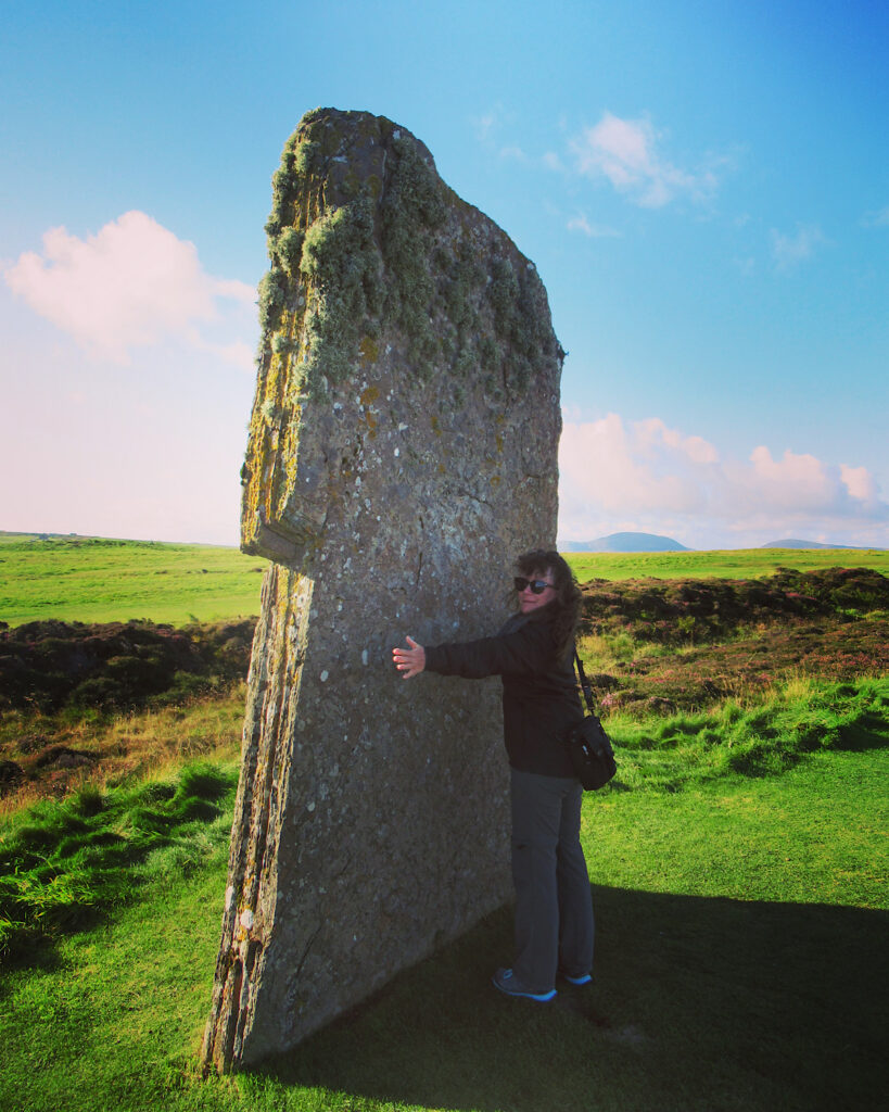 Woman in black coat and grey pants standing in front of massive, upright, stone slab with her arms outstretched and not coming close to reaching width of the stone.