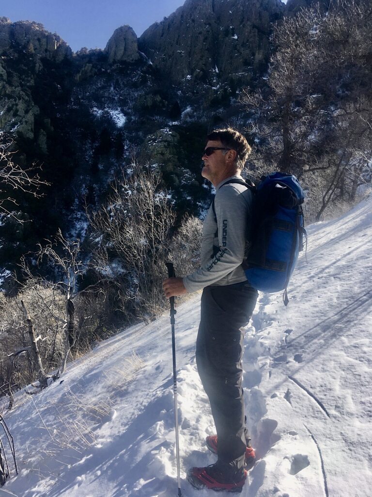 Man in long sleeve gray shirt and dark grey pants with blue backpack and hiking boots and a pole looking out from snow-covered slope.