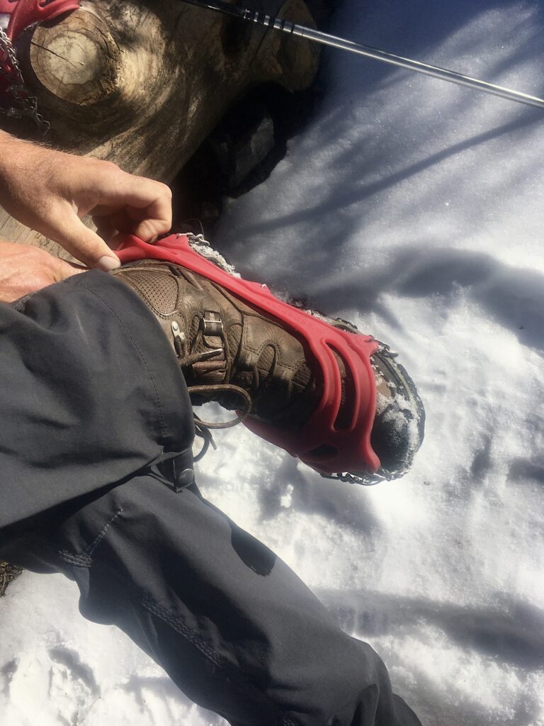 Man putting red rubber traction grips on leather boots in snow.