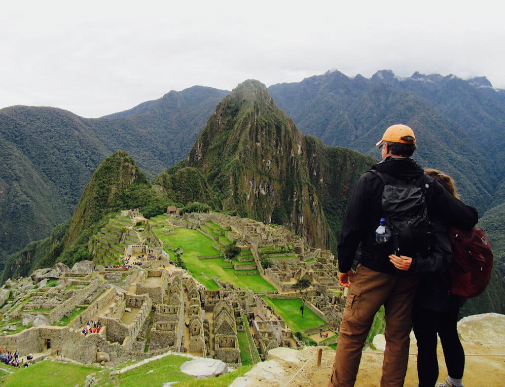 Man and woman in long pants and coats facing out over stone ruins on rugged green mountain hillsides.