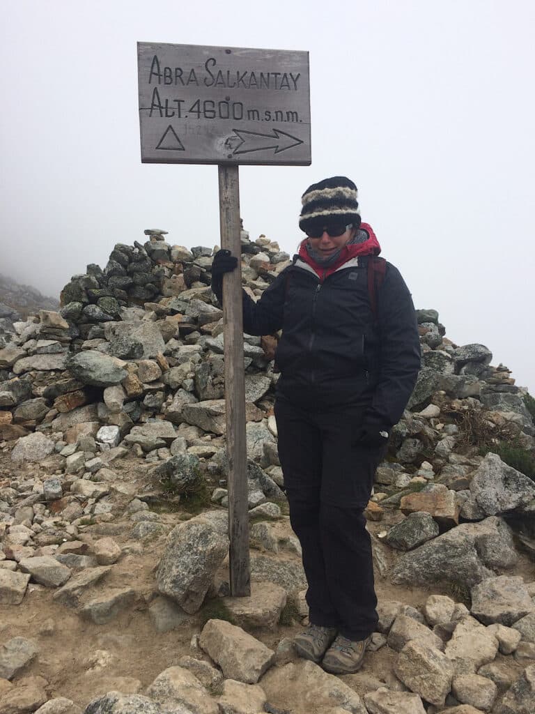 Woman in black rain jacket and black pants with white and black toque standing by wooden signpost reading: Abra Salkantay Alt. 4600 m.s.n.m.