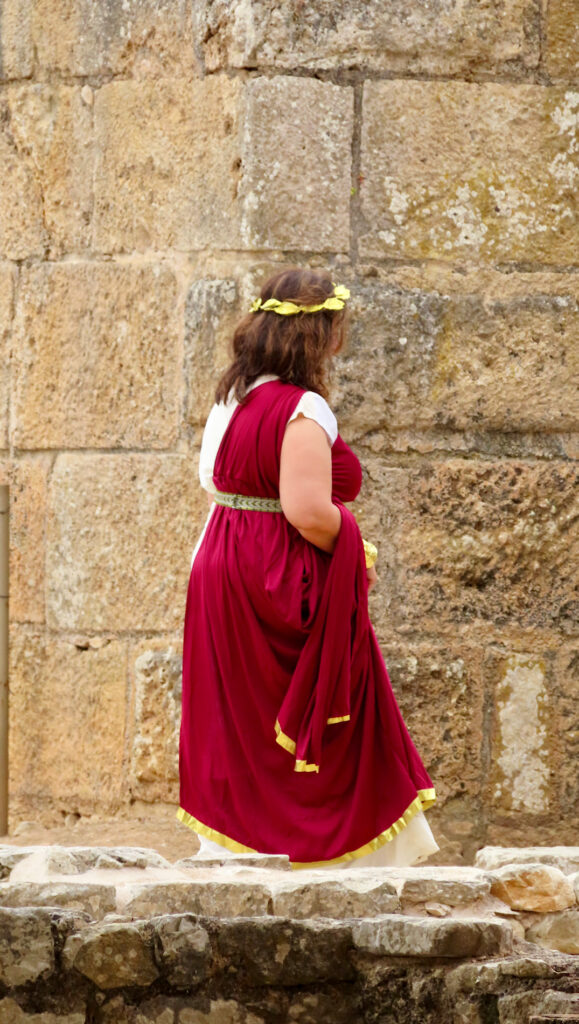 Woman in deep red toga with white below, gold leaf headwreath and gold belt walking by thick stone wall.