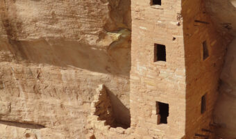 Multi-storey high square stone building with 3 windows on front built against high sandstone cliff face.