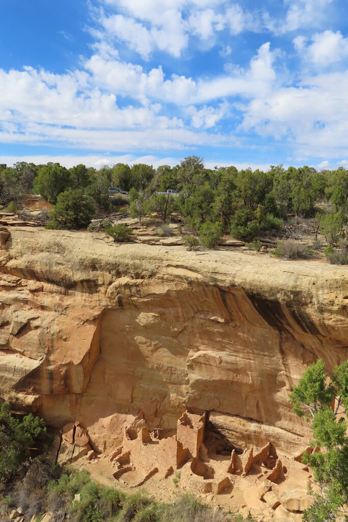 Crumbling ruin beneath high reddish brown sandstone cliff topped with green vegetation.