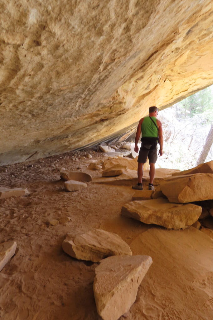 Man in green tank top and shorts walking through overhanging sandstone alcove with chunks of stone on floor of cave.