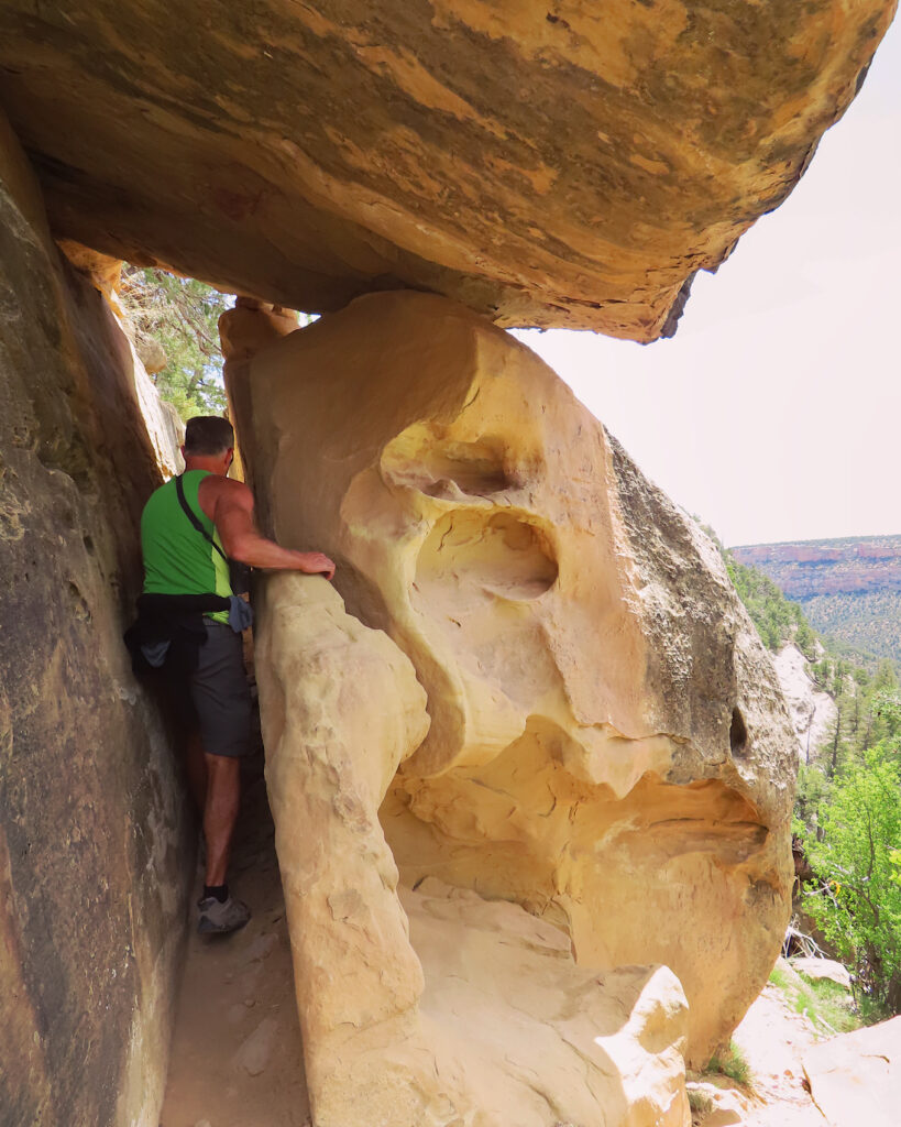 Man in green shirt passing between narrow gap of stone with large stone covering gap above his head.