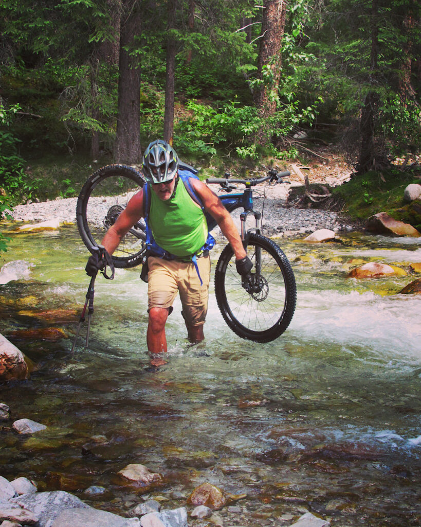 Man in brown shorts and green short sleeve shirt carrying bike on shoulders as he wades across large creek.