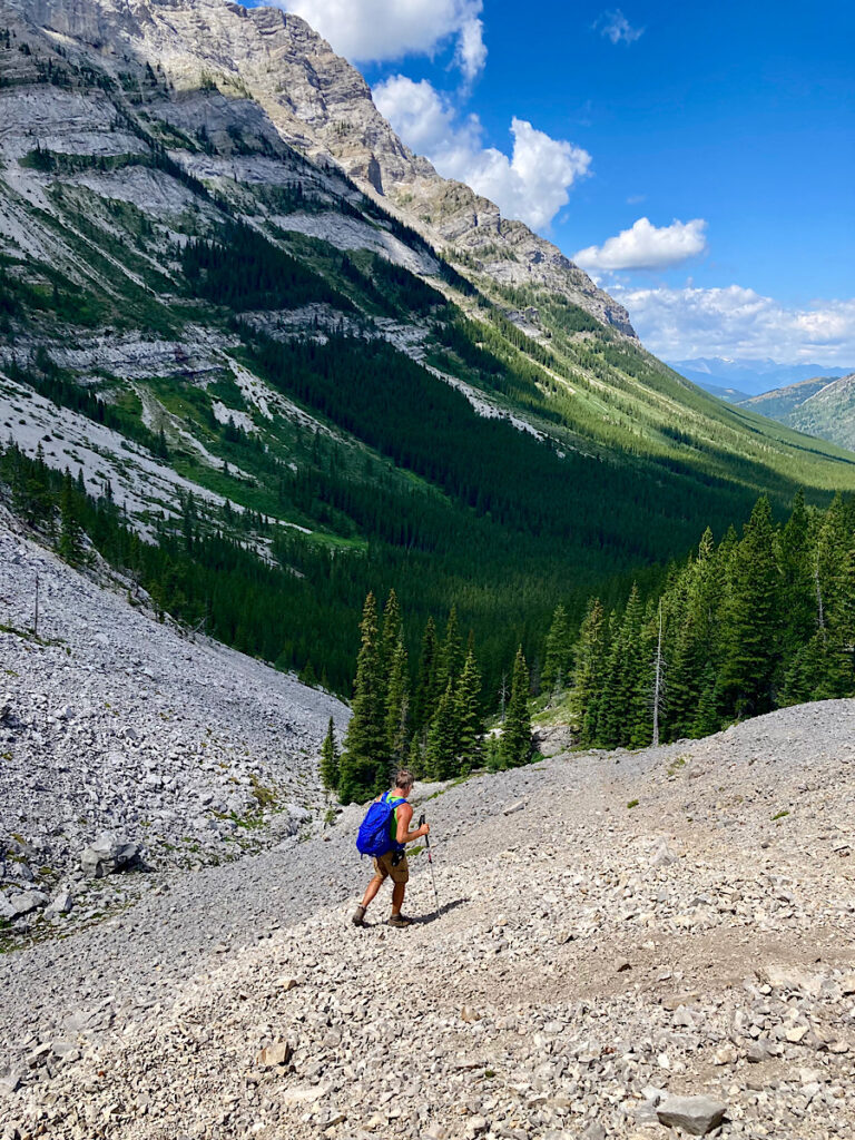 Male hiker on scree slope above thickly forested valley beneath blue sky.