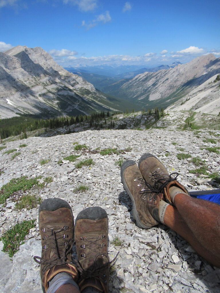 Outstretched legs of hikers showing two pairs of boots and valley and mountain below.