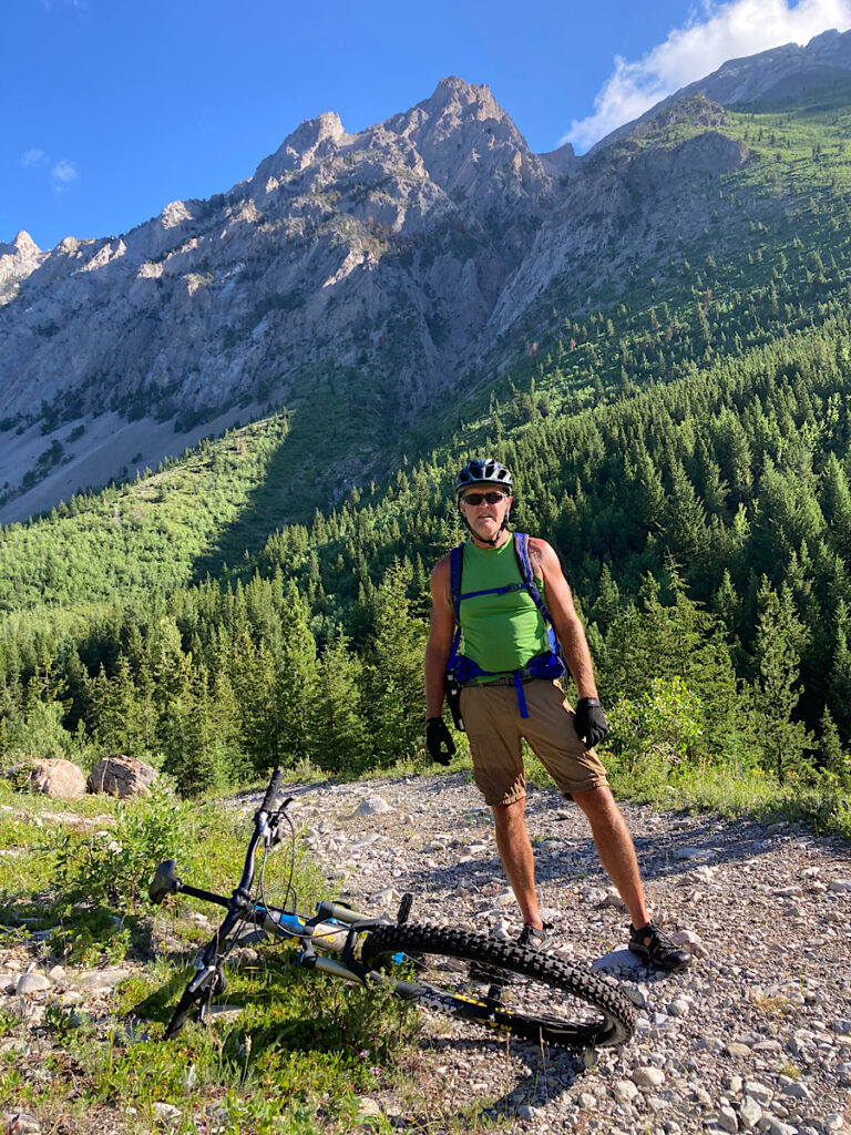 Man in green short sleeve shirt and brown shorts standing on gravel area above treed valley with tall mountains in distance under blue sky.