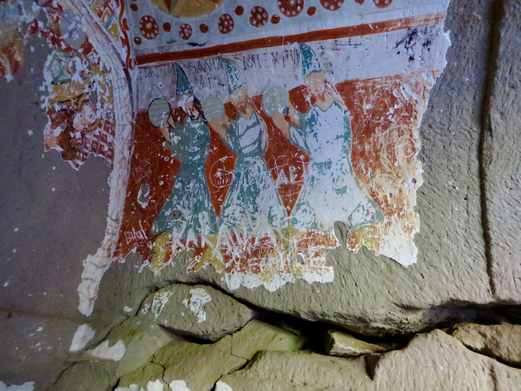 Old stone wall with peeling painting of three men in blue cloaks with red background.