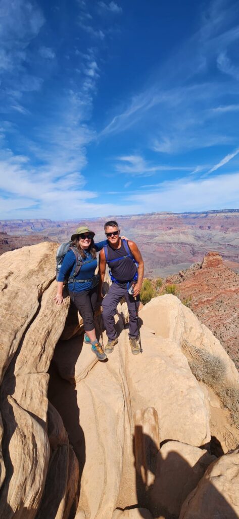 Man and woman hikers standing on brown rock overlooking deep reddish canyon under blue sky.